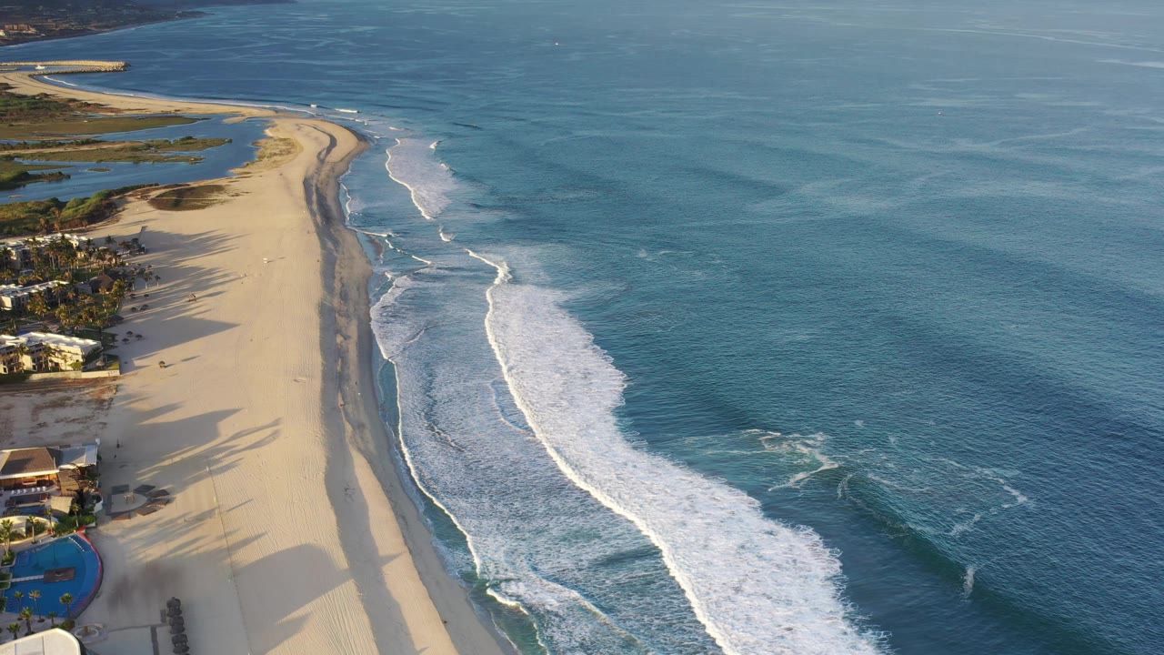 The aerial view continues along the Los Cabos shoreline, showing more open water on the right, surf curving toward the sand, and residential buildings along the left edge with no visible people.