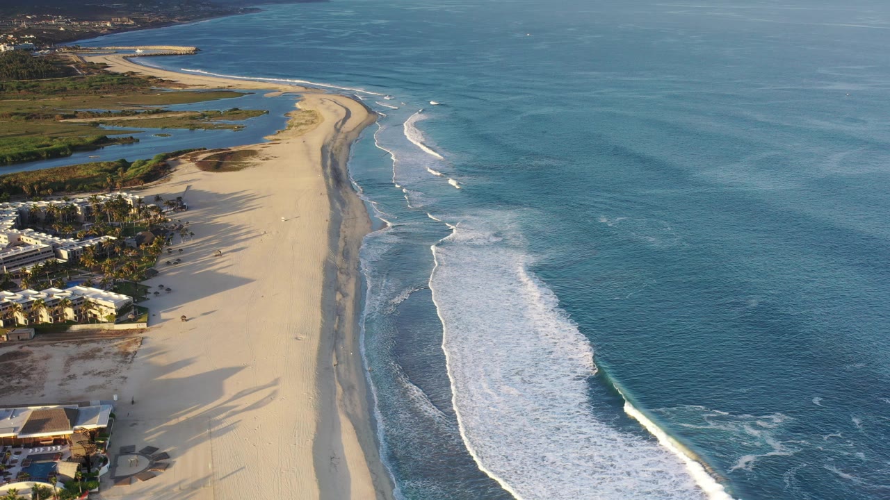 Aerial view over a Los Cabos beach with oceanfront buildings at lower left, a long strip of sand, breaking surf to the right, and no visible people.