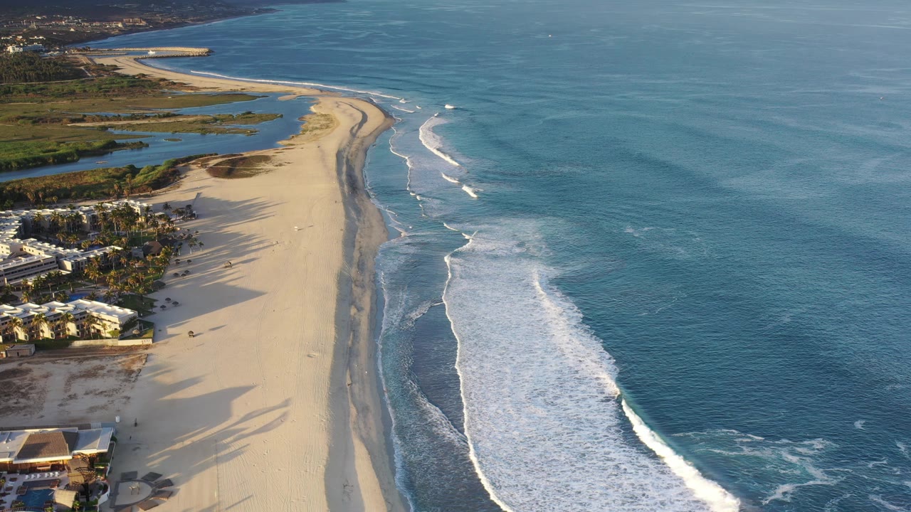 Wide aerial coastline with beach, ocean, surf, and resort-style buildings.