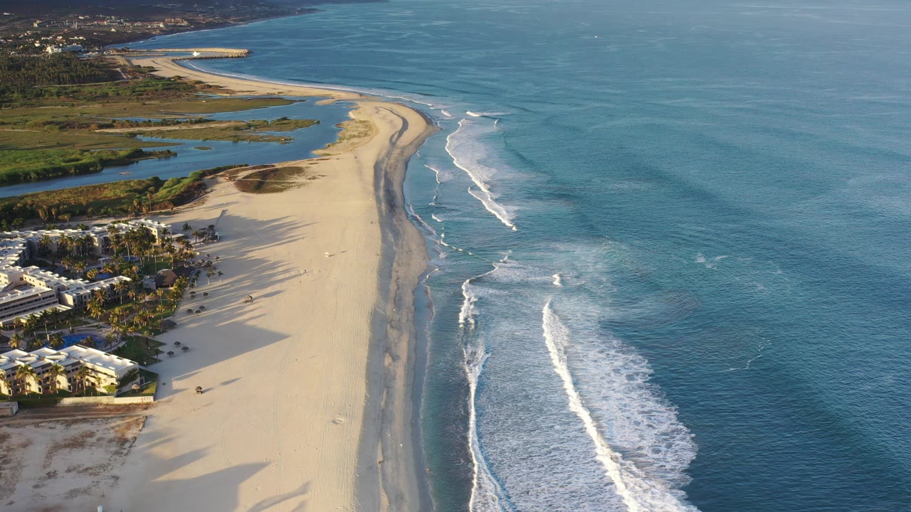 High aerial view over a Los Cabos sandy beach with blue ocean on the right, white surf bands near shore, inland water at upper left, beachfront buildings at lower left, and no visible people.