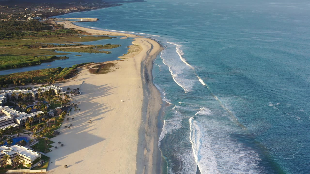 Aerial view over a Los Cabos beach with white surf on the right, lagoon water inland, beachfront buildings at the lower left, distant mountains, and no visible people.