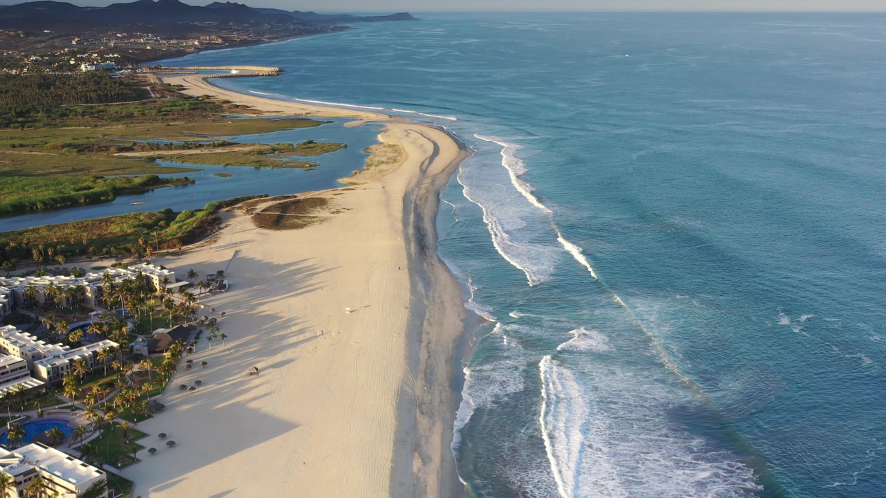 Aerial coastline with open ocean, surf bands, sandy beach, wetlands, and mountains.
