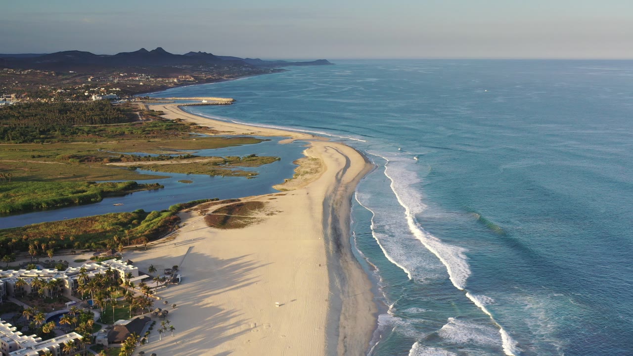 Aerial view over Los Cabos, Mexico, with a curving sand beach, blue ocean surf, lagoon water, low buildings and palms in the lower foreground, distant hills, and tiny possible people on the beach.