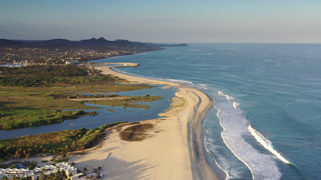 Aerial view over Los Cabos with a curved sandy beach between blue ocean waves and green-brown wetlands, distant hills, coastal buildings, and no visible people.