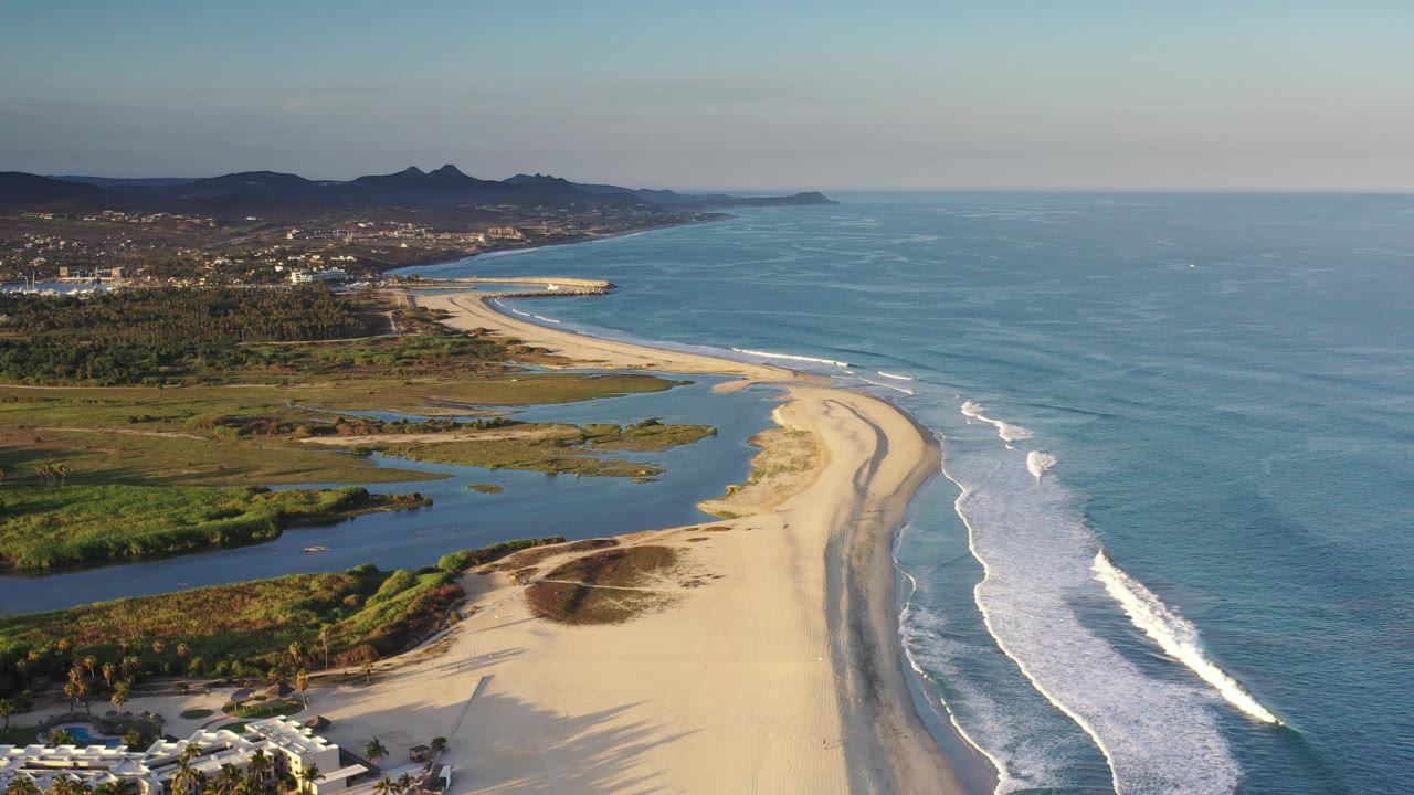 Wide aerial frame of Los Cabos coastline showing the sandbar curving along the surf, wetland pools inland, low hills on the horizon, and no visible people.