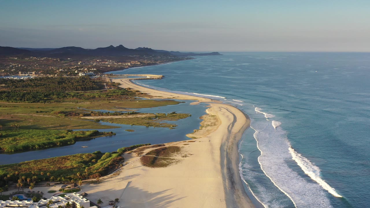 Aerial beach curve with waves, wetlands, distant town, and mountains.