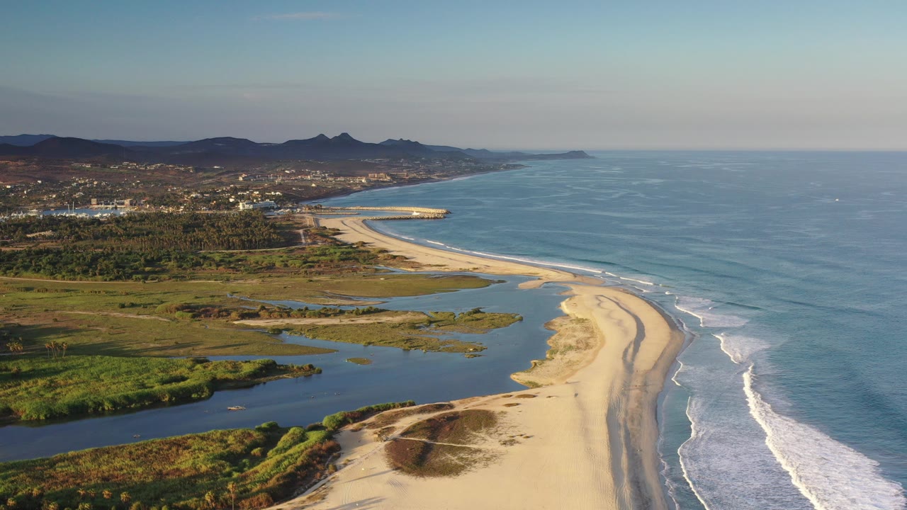 Opening aerial frame over the Los Cabos, Mexico coastline, with a curved sand beach, dark lagoon water, blue ocean surf, distant hills, low buildings, and no visible people.
