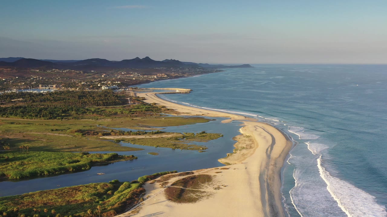 Aerial view along the Los Cabos, Mexico shoreline, showing the sandbar and beach slightly farther into frame, with ocean waves to the right, lagoon water to the left, and no visible people.