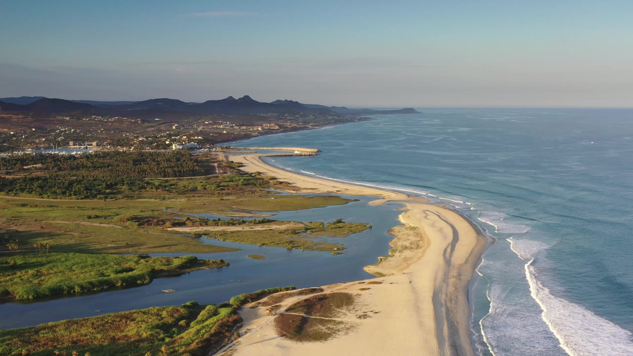 Wide drone-like coastal frame with beach, surf, lagoon pools, town, hills, and open sky.
