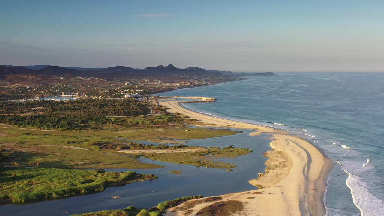 Opening aerial frame over the Los Cabos coastline, with a sandy beach curving along blue surf, lagoon water in the foreground, inland buildings, distant mountains, and no visible people.