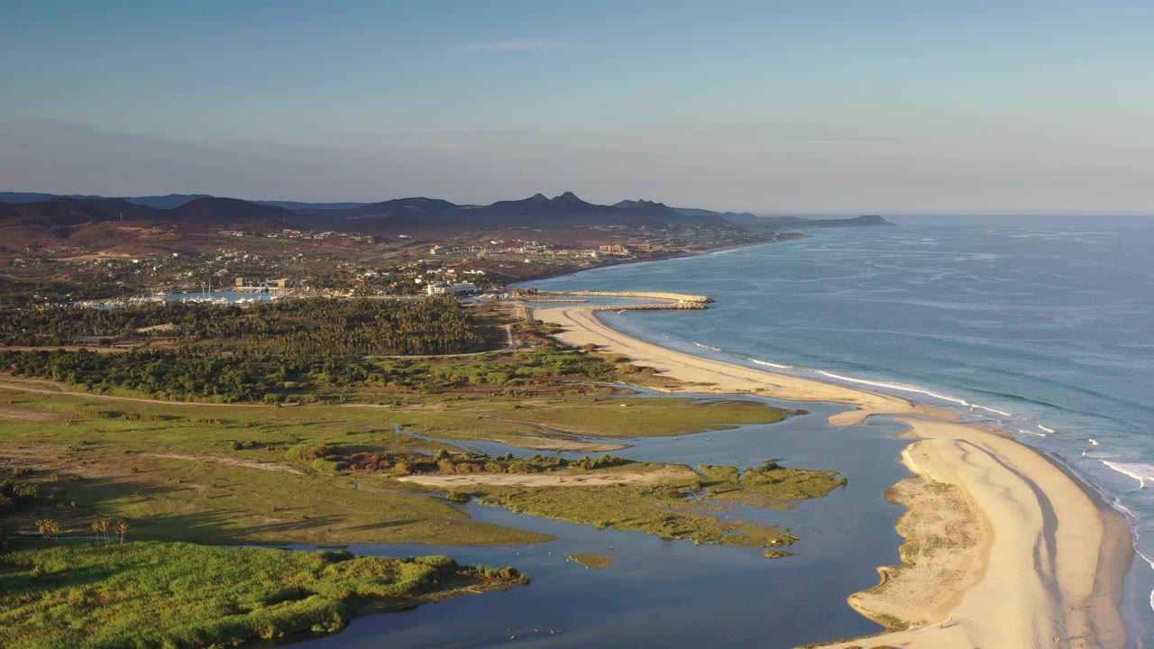 Aerial view in Los Cabos, Mexico with lagoon water in the foreground, a curving sandy beach beside blue ocean, inland trees, low hills, scattered buildings, and no visible people.