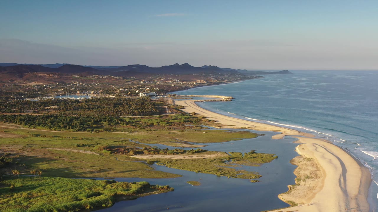 Aerial Los Cabos coastline showing lagoon channels across the foreground, pale sand meeting small ocean waves, a green coastal strip, inland buildings, distant hills, and no visible people.
