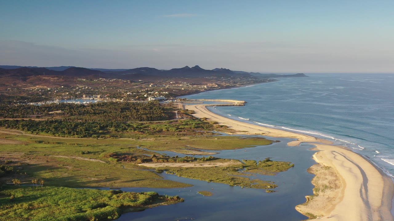 Wide aerial frame in Los Cabos, Mexico with the ocean filling the right side, wave lines touching the beach, lagoon and marshy vegetation below, inland development, hills, and no visible people.