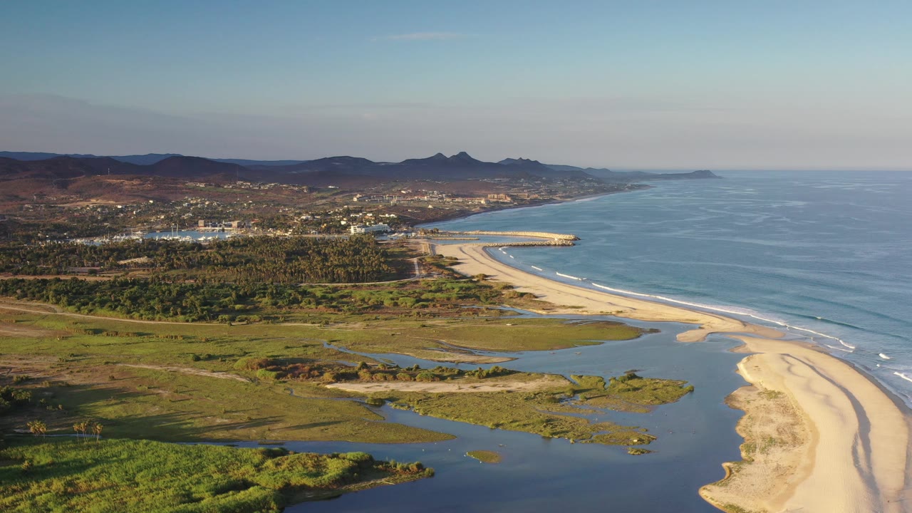 Coastal aerial with lagoon foreground, sandy shore, breaking waves, trees, town, and hills.