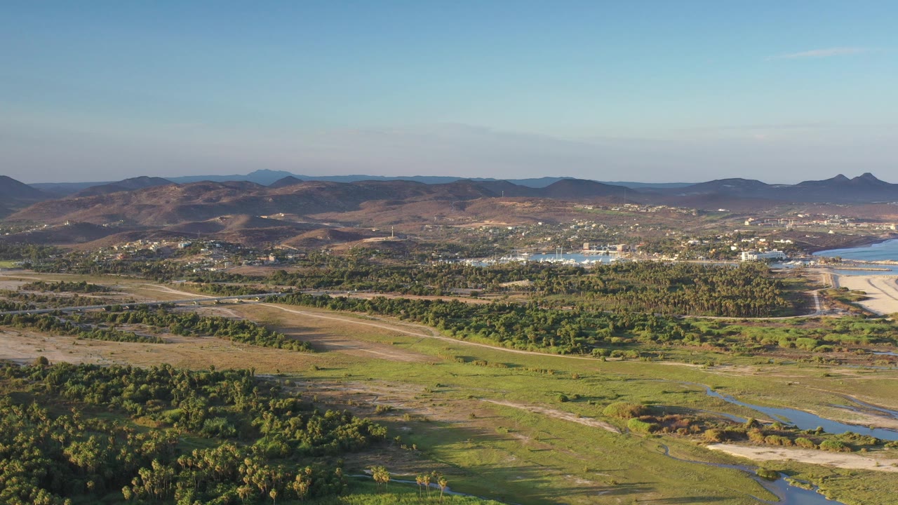 Opening aerial frame over Los Cabos, Mexico, with green foreground vegetation, narrow water channels, distant buildings, mountain ridges, clear blue sky, and no visible people.