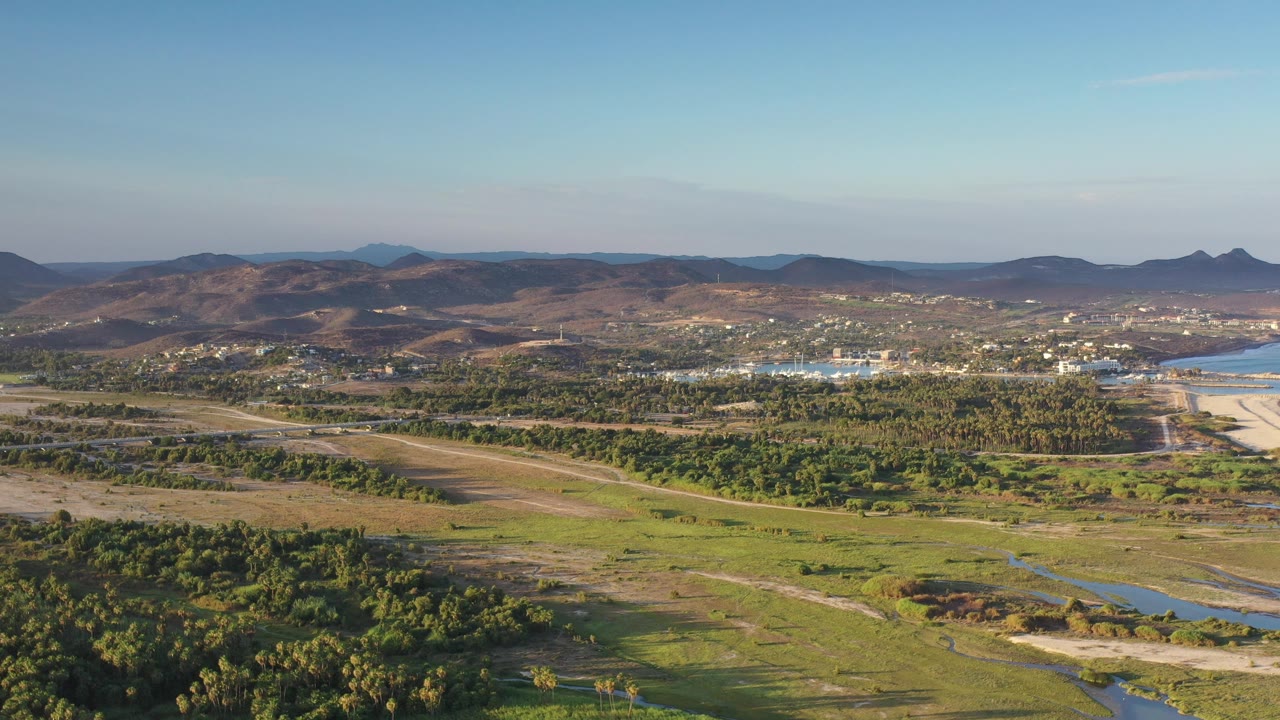 Broad aerial view across trees, open land, water, distant buildings, and mountain horizon.