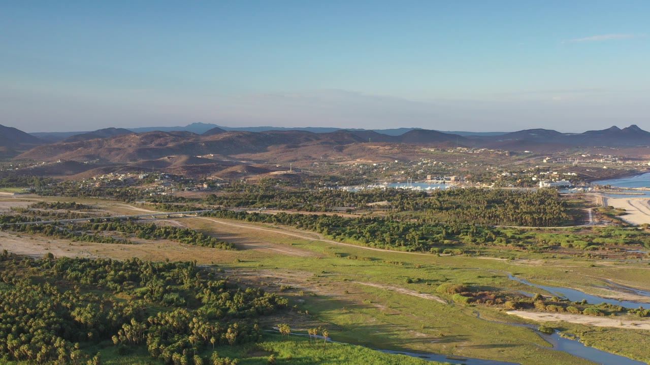 Wide aerial view in Los Cabos, Mexico, with green grassland and forest in the foreground, pale shoreline and water to the right, distant brown mountains, and no visible people.