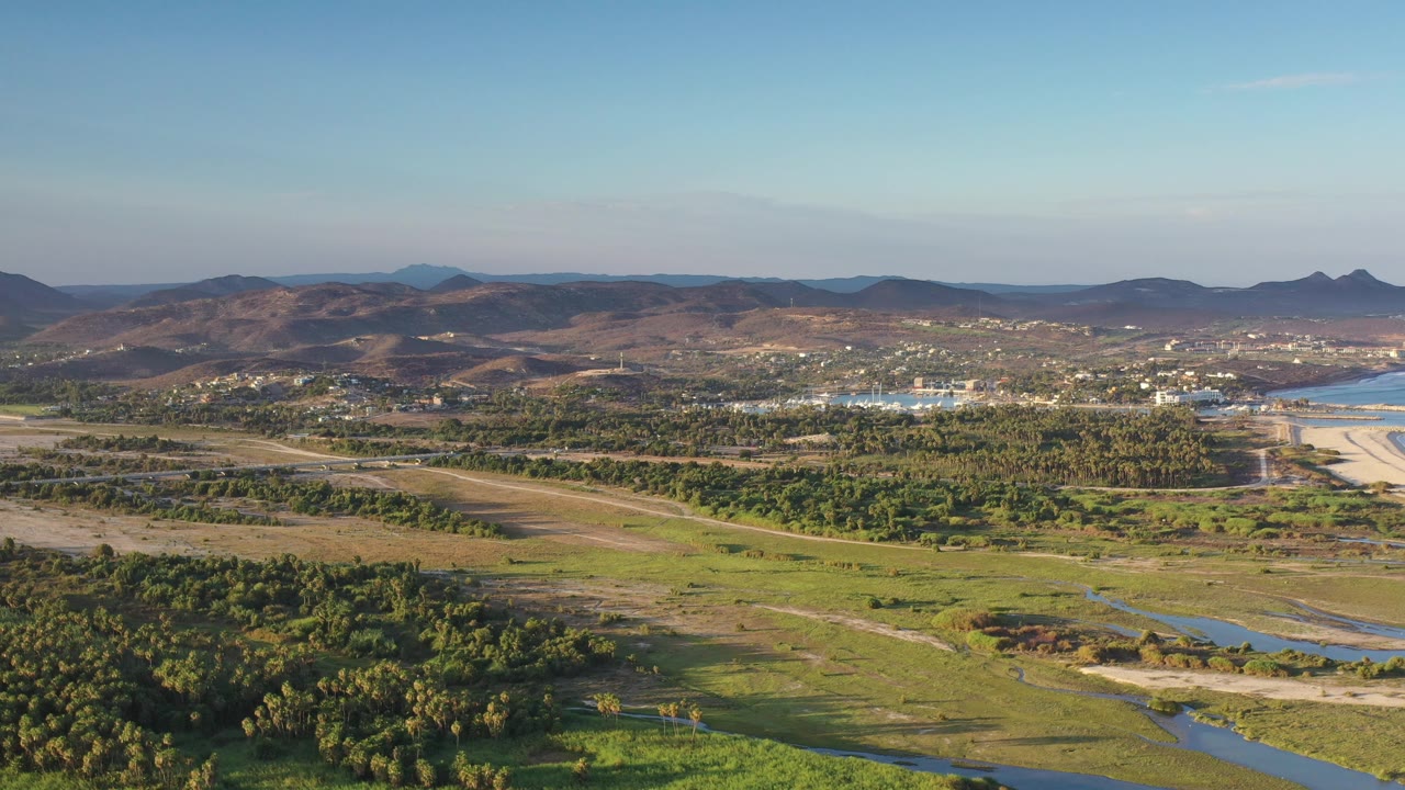 Aerial view across green lowland toward brown hills and a bright water edge.