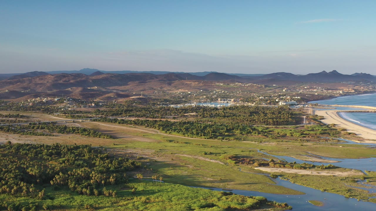 Opening frame of Los Cabos, Mexico from above, with green wetland channels in front, a pale beach curving beside blue water, dry mountains in back, and no visible people.