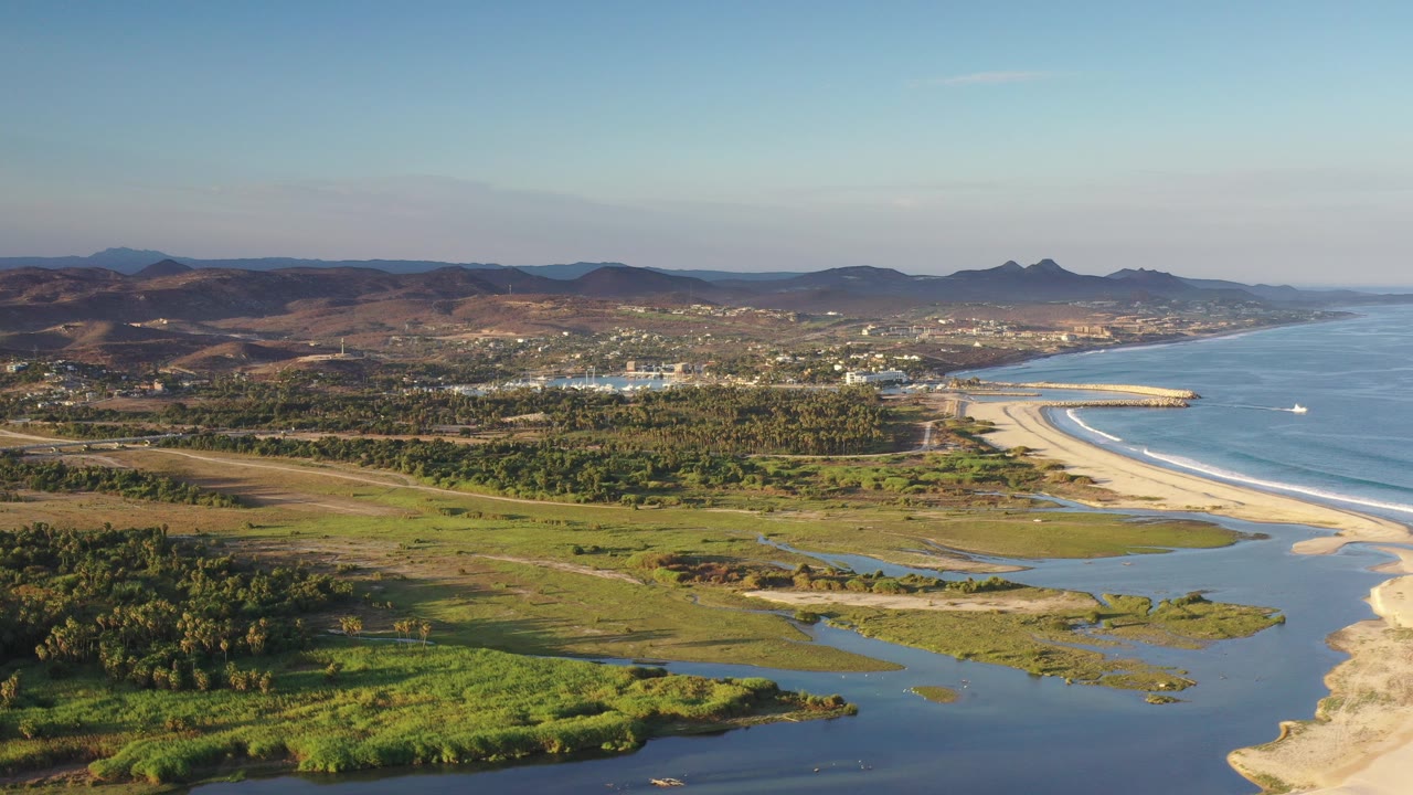 Aerial view over Los Cabos with lagoon channels in the foreground, a curved beach and ocean to the right, distant buildings, mountains on the horizon, and no visible people.