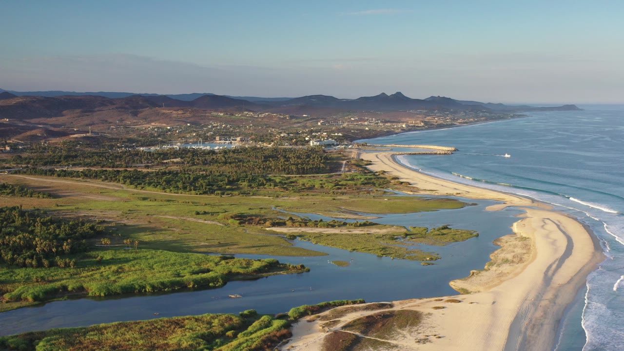 Opening aerial frame over Los Cabos, Mexico, showing a sandy beach curve, green lagoon channels, blue ocean surf, distant town, brown mountains, and no visible people.