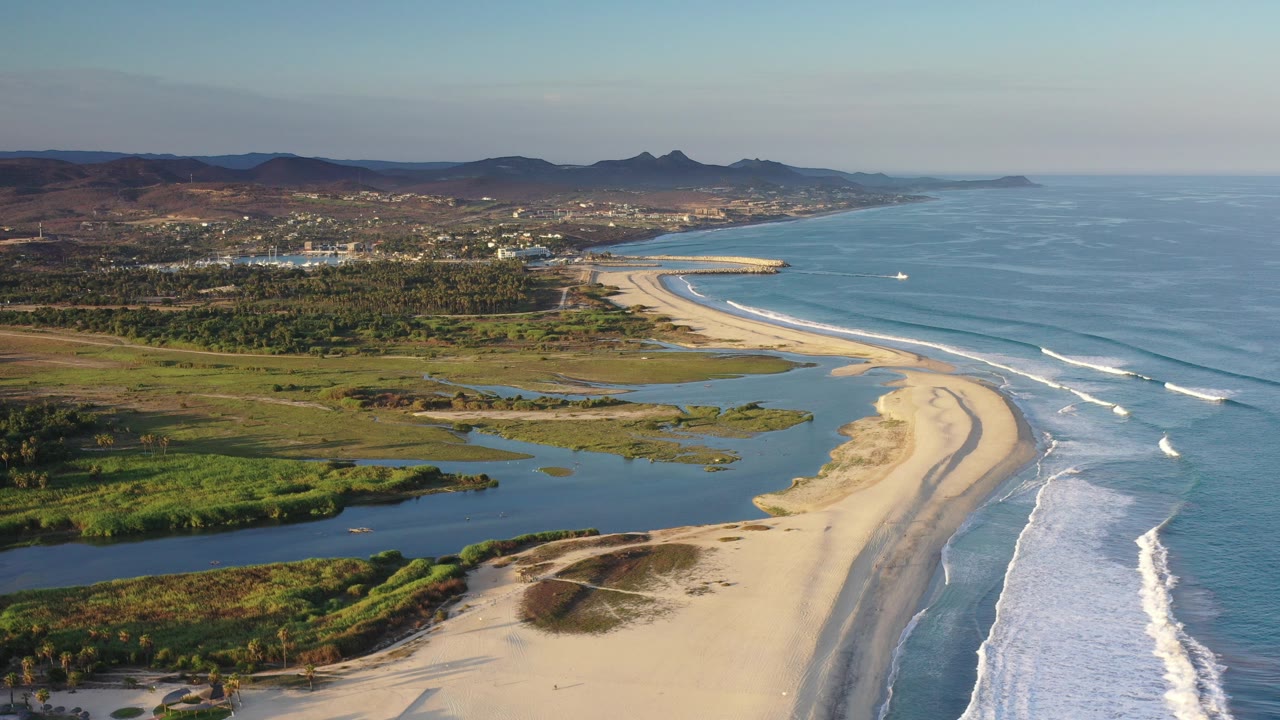 Aerial view of sandy coastline, blue ocean waves, lagoon water, green wetlands, town, and hills.