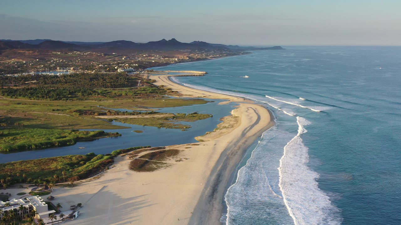 Aerial view of curved sandy beach, surf, lagoons, hills, and ocean.