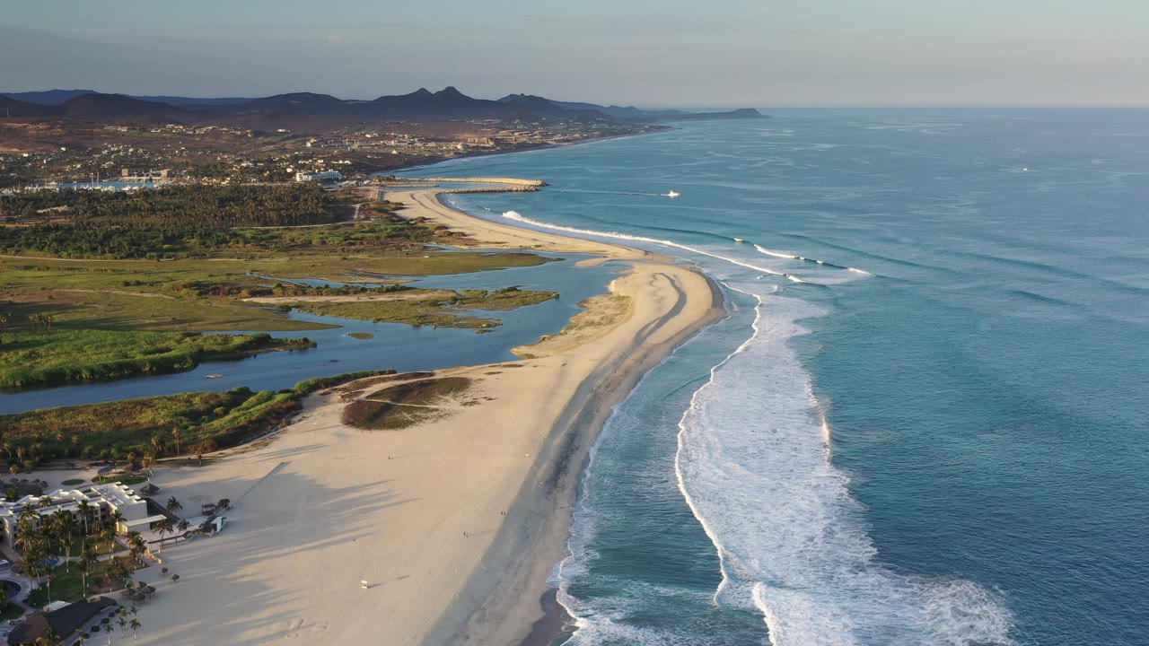 Aerial view over a Los Cabos beach with white surf curving along sand, lagoon water inland, distant town and hills, and no visible people.