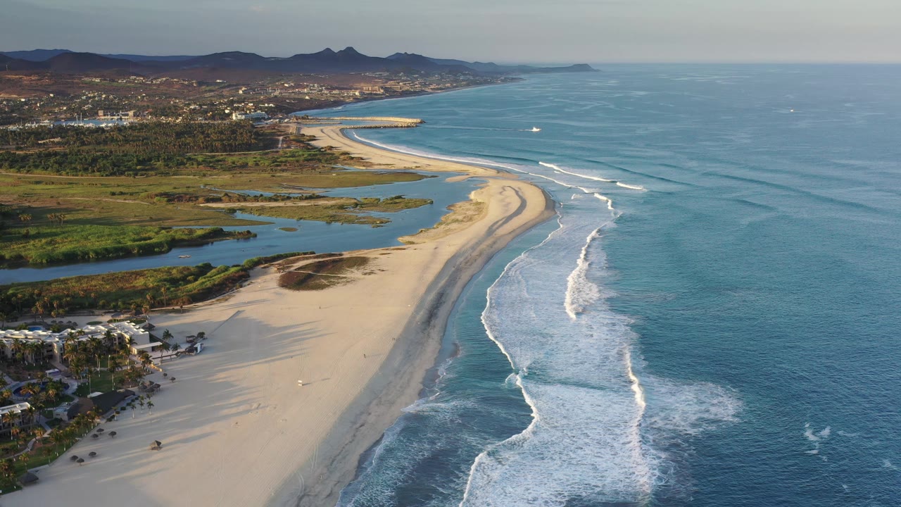 Elevated Los Cabos coastal view with the curving sandbar centered, white waves breaking closer to shore, lagoon-like water inland, palms and buildings below, and no visible people.