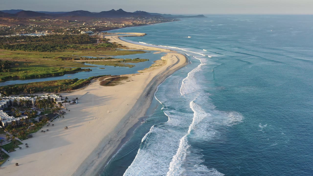 Aerial frame from Los Cabos, Mexico, with a long pale sandbar, angled bands of breaking surf, blue water to the right, green inland terrain to the left, and small beach figures in the distance.