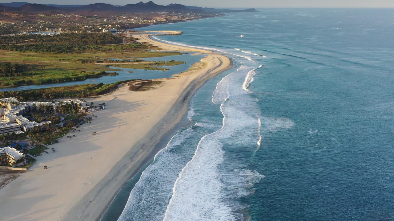 A high aerial view over a Los Cabos beach shows white surf bands meeting pale sand, blue ocean to the right, inland lagoons and buildings to the left, distant hills, and no visible people.