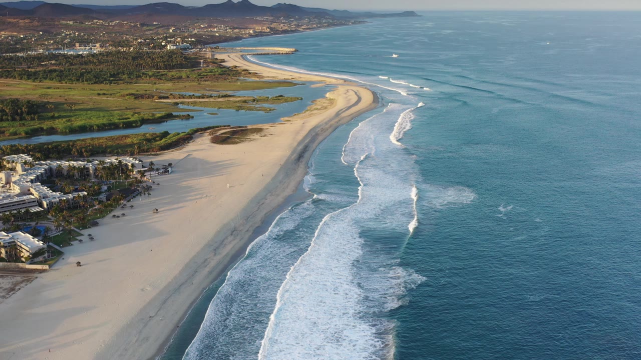 Drone-like view of beach, ocean surf, lagoons, and distant hills.
