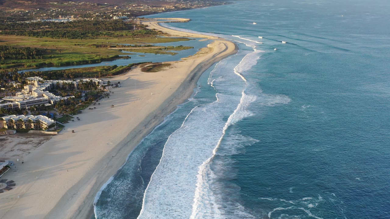 Aerial view of a sandy beach, surf, blue ocean, coastal buildings, and inland greenery.