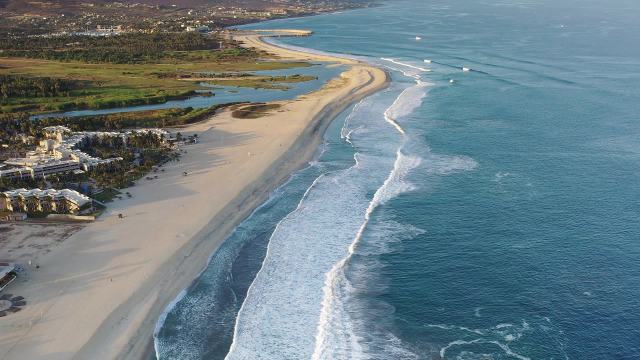 Aerial Los Cabos shoreline frame with long white wave lines advancing toward the beach, resort-like coastal buildings at lower left, green inland water features, small boats offshore, and no visible people.