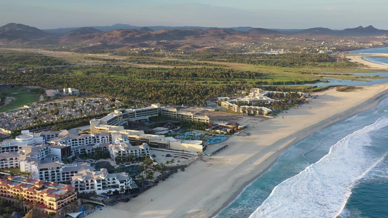 Aerial view in Los Cabos showing resort roofs and pools near the lower edge, beach and foamy surf along the right side, palm-covered land behind the hotels, hazy hills on the horizon, and no visible people.