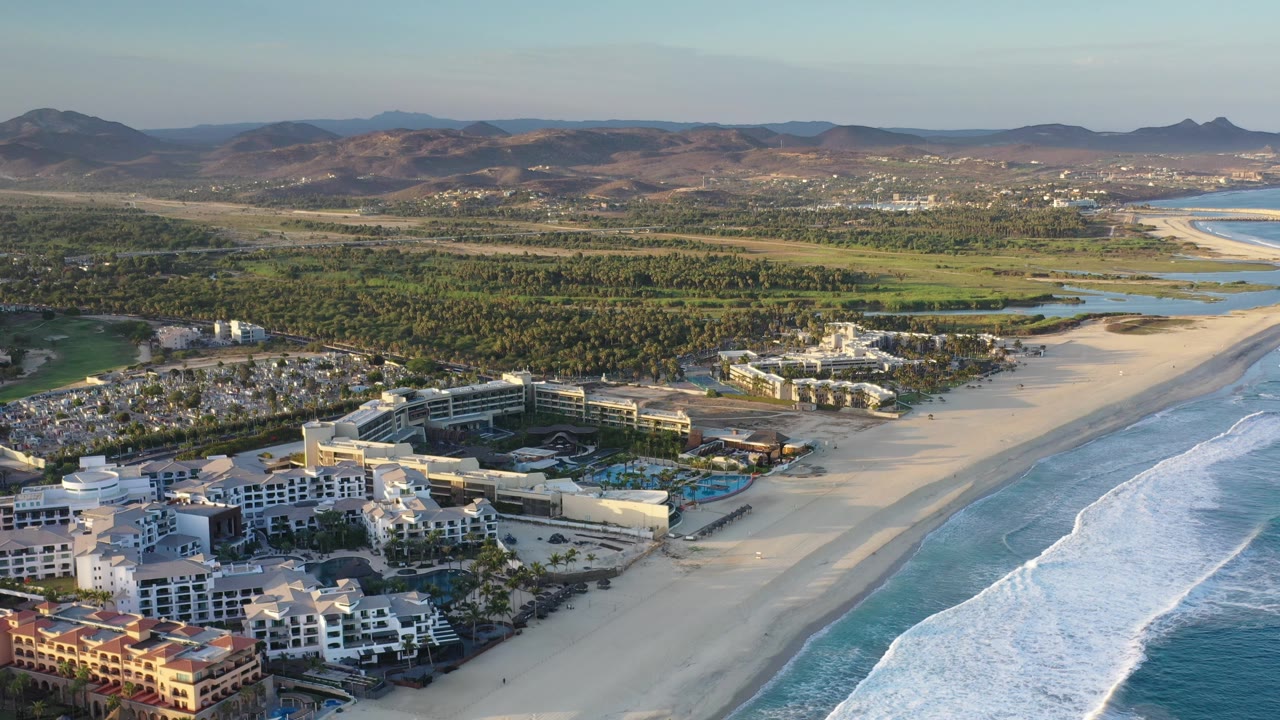 Wide Los Cabos aerial with the resort cluster across the lower left, empty-looking sand stretching up the coast, white surf bands at frame right, green lowlands inland, and no visible people.