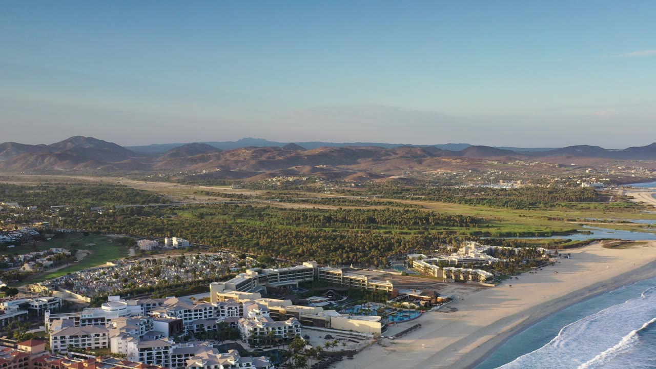 Wide aerial view of beachfront buildings, surf, green inland terrain, and low mountains.