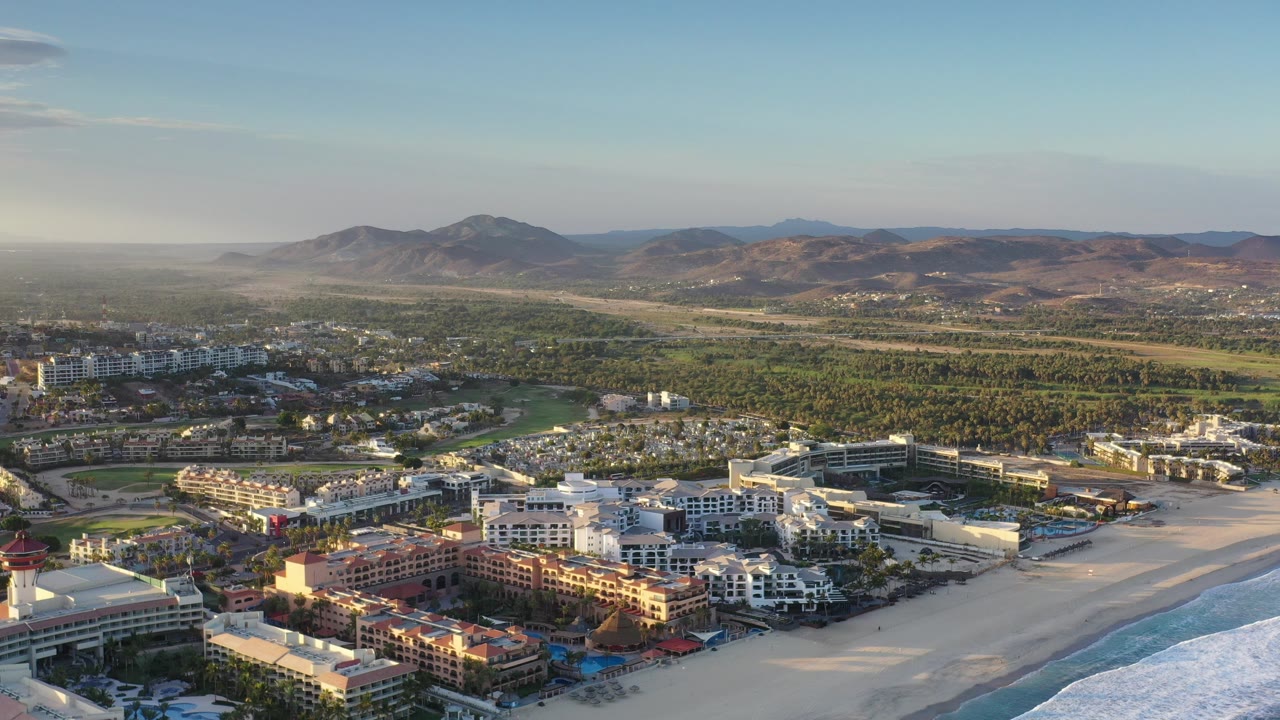 Opening aerial frame over Los Cabos, Mexico with beachfront hotel blocks, pale beach, blue surf at the right edge, low city development, distant hills, and no visible people.