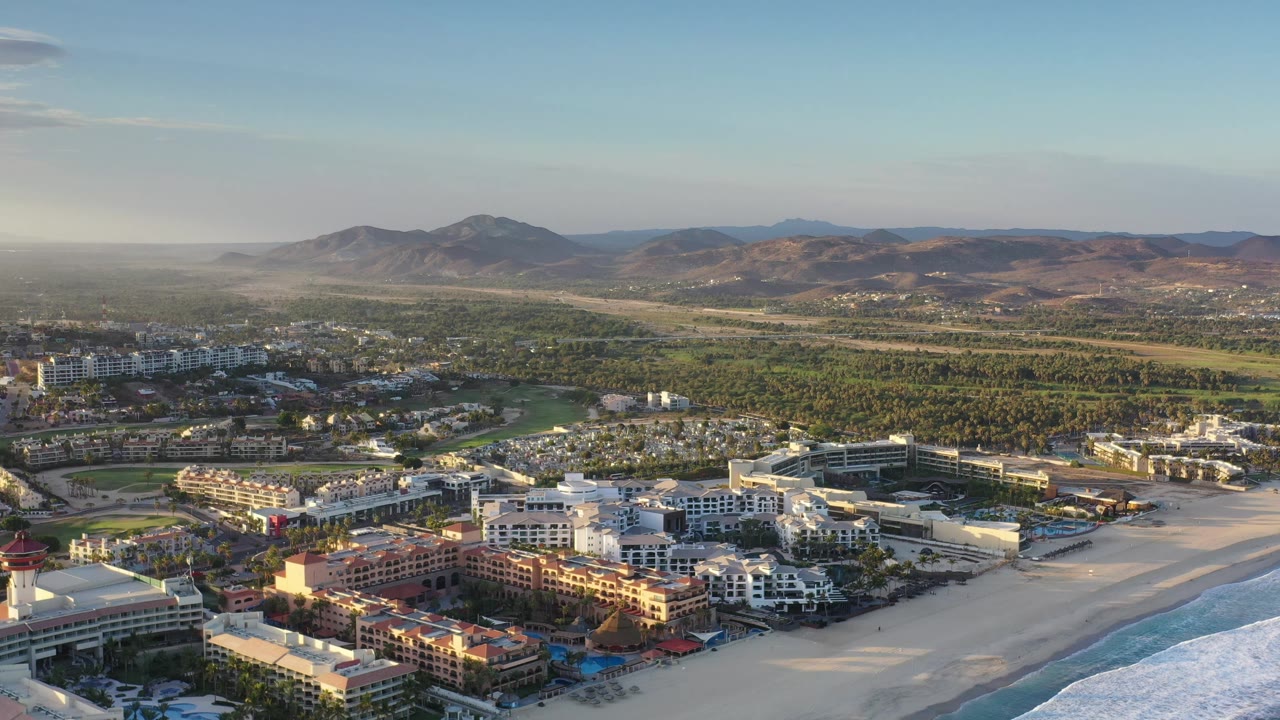 Aerial view in Los Cabos, Mexico with sunlit resort buildings in the lower frame, surf along the right, spread-out city blocks, green areas, hazy distance, and no visible people.
