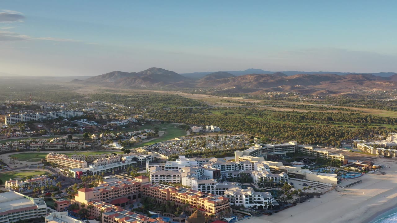 Los Cabos aerial view with resort blocks and parking lots at the bottom of frame, green terrain in the midground, hazy desert hills in the distance, and no visible people.