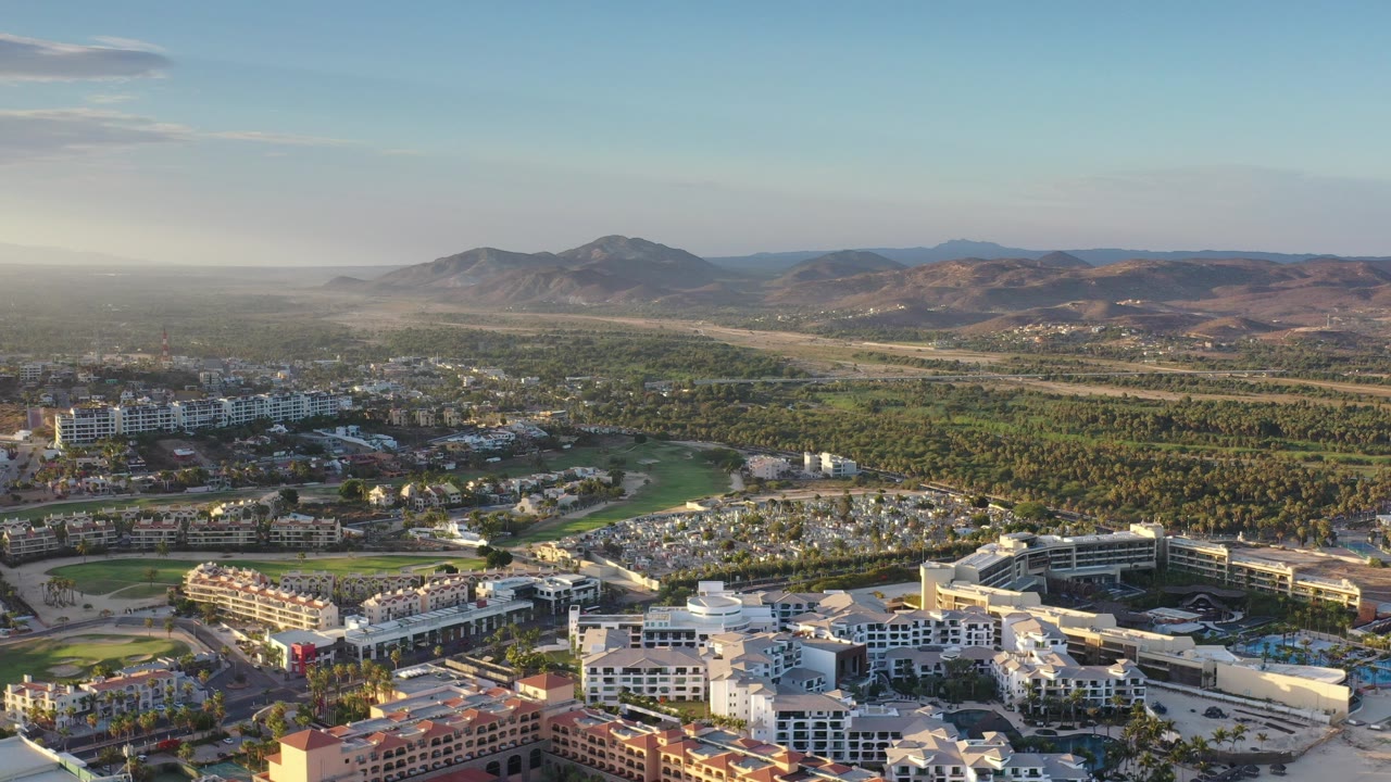 Opening frame of a Los Cabos aerial view with low-rise buildings in the foreground, palms and green developed land in the middle distance, hazy mountains on the horizon, and no visible people.