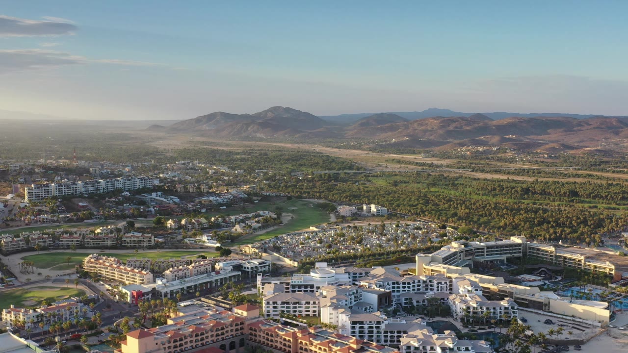 At 0:01, the Los Cabos aerial holds on a broad city-and-landscape layout with white buildings, palms, green fairway-like areas, dry hills, distant mountains, and no visible people.