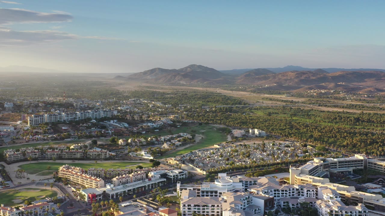 Aerial view over buildings, golf greens, trees, and mountains.