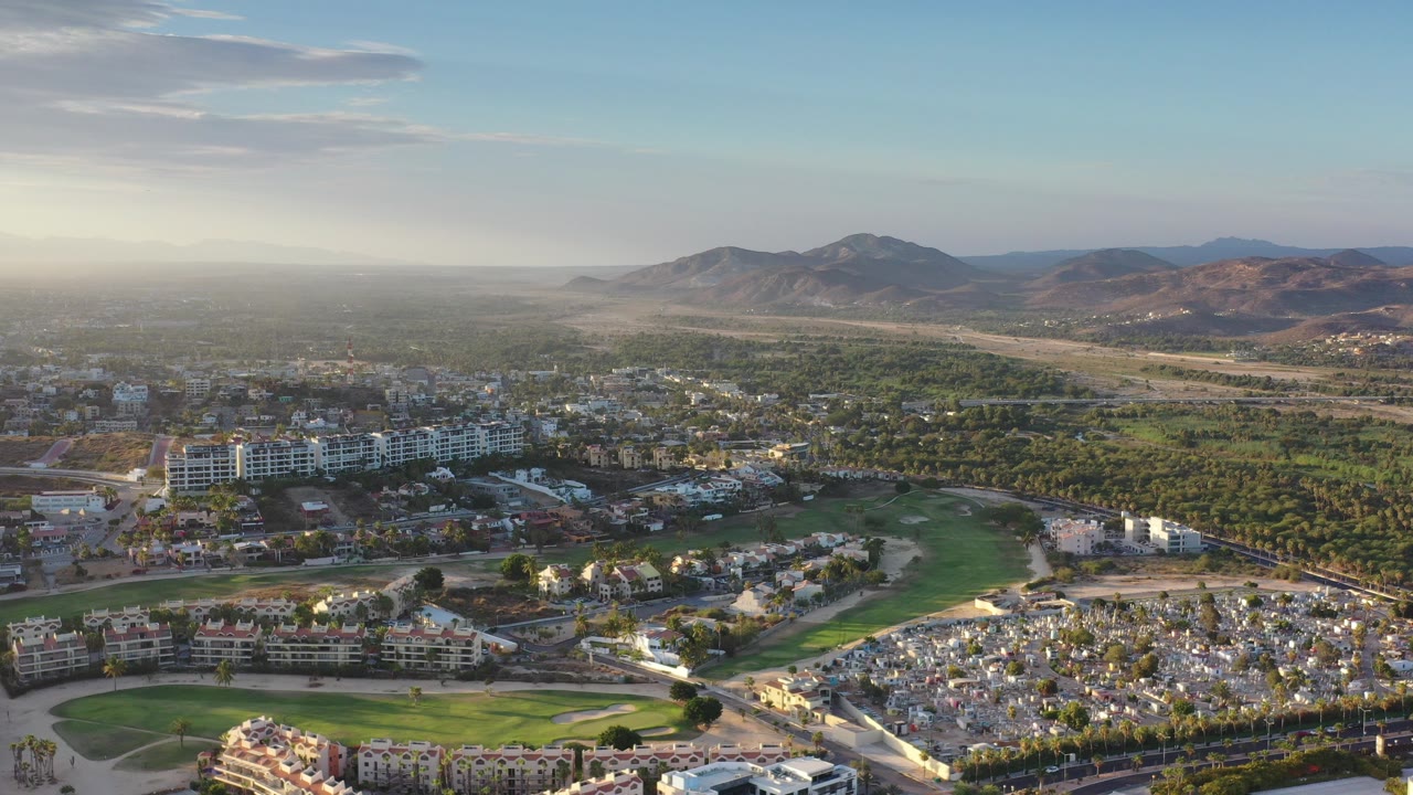 Opening aerial frame over Los Cabos, Mexico, with golf greens and low-rise buildings in the foreground, tree cover across the middle distance, mountains on the horizon, and no visible people.