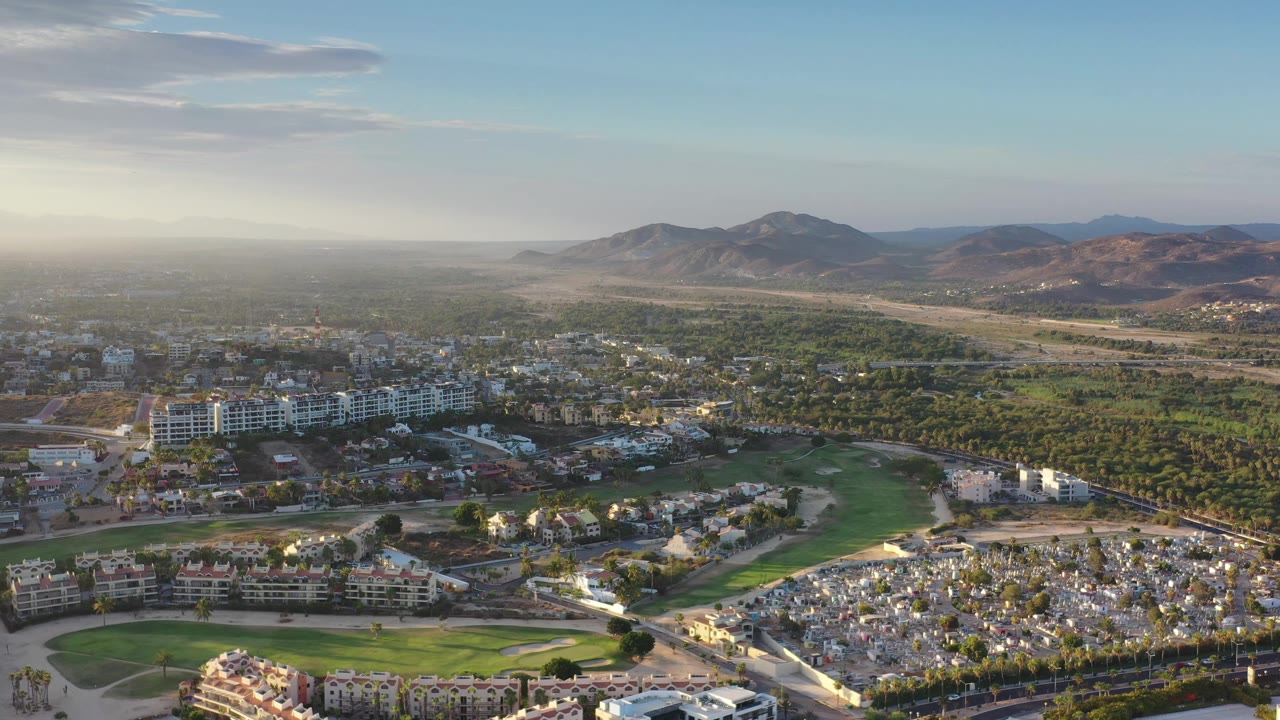 Aerial Los Cabos frame with landscaped green areas and clustered buildings below, a tree-filled plain beyond, dry mountains in the background, and no visible people.