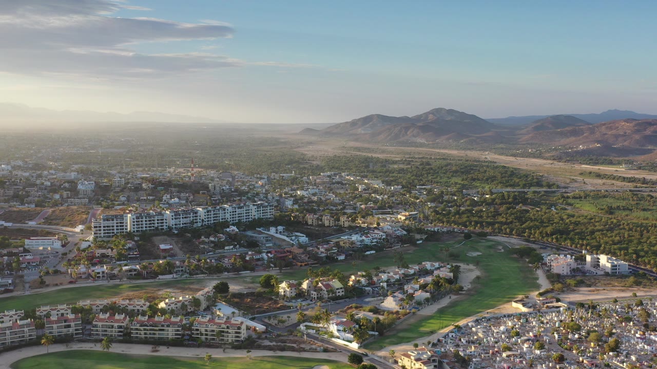 Wide aerial view of Los Cabos rooftops, green fairway, desert flats, and distant hills.