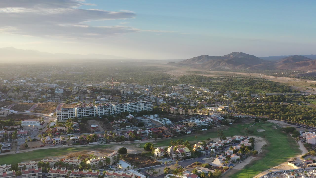 Aerial view over Los Cabos, Mexico, showing white mid-rise buildings, residential blocks, a green open area, tree cover, hazy hills, and no visible people.