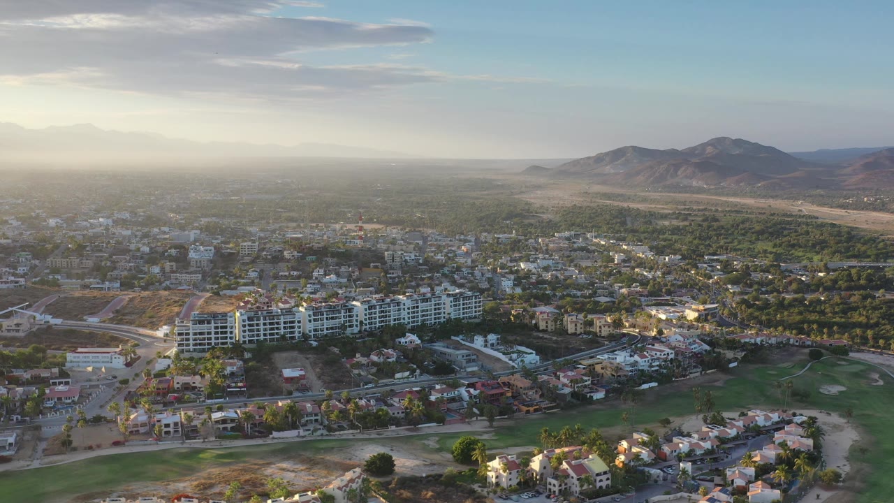 Aerial frame over Los Cabos with the white building and nearby homes slightly more centered, backed by green vegetation, mountains, pale sky, and no visible people.