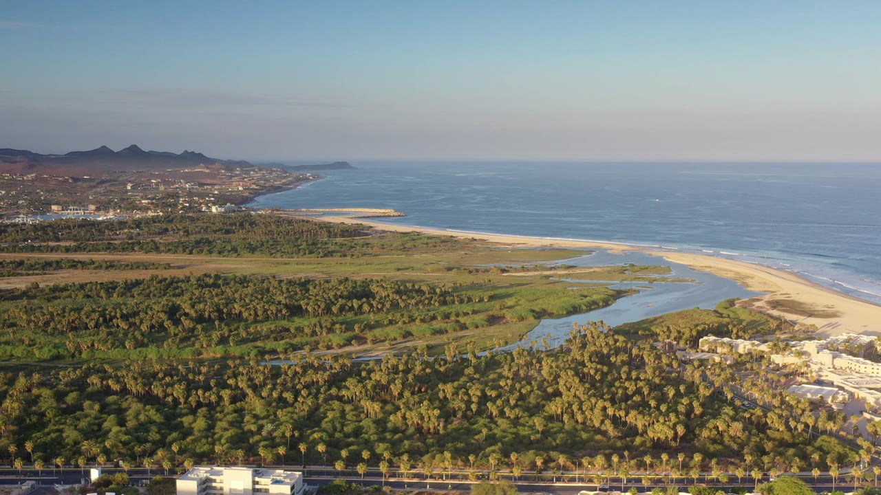 Wide aerial frame over Los Cabos, Mexico, with blue ocean to the right, a pale beach and lagoon channel in the middle, palm-covered land below, distant hills, and no visible people.