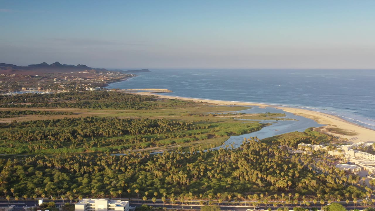Aerial view in Los Cabos, Mexico, showing palm groves in the lower frame, a curving lagoon and sandbar, the ocean horizon, distant buildings along the coast, and no visible people.
